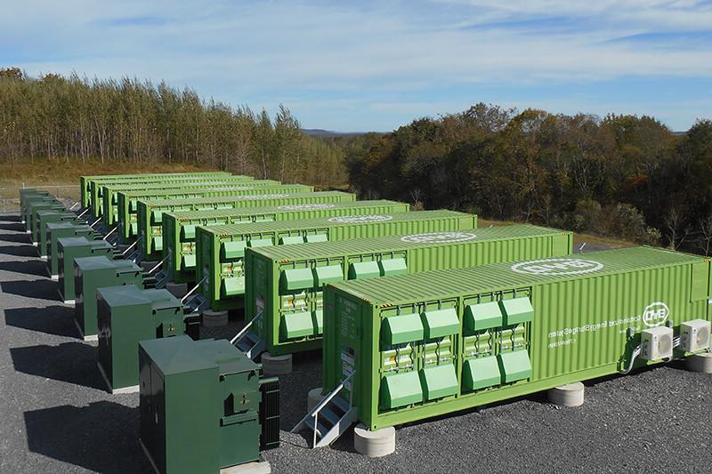 A line of large green corrugated metal containers, used for 能源 battery storage. They are on an asphalt surface, surrounded by trees and with a blue sky above